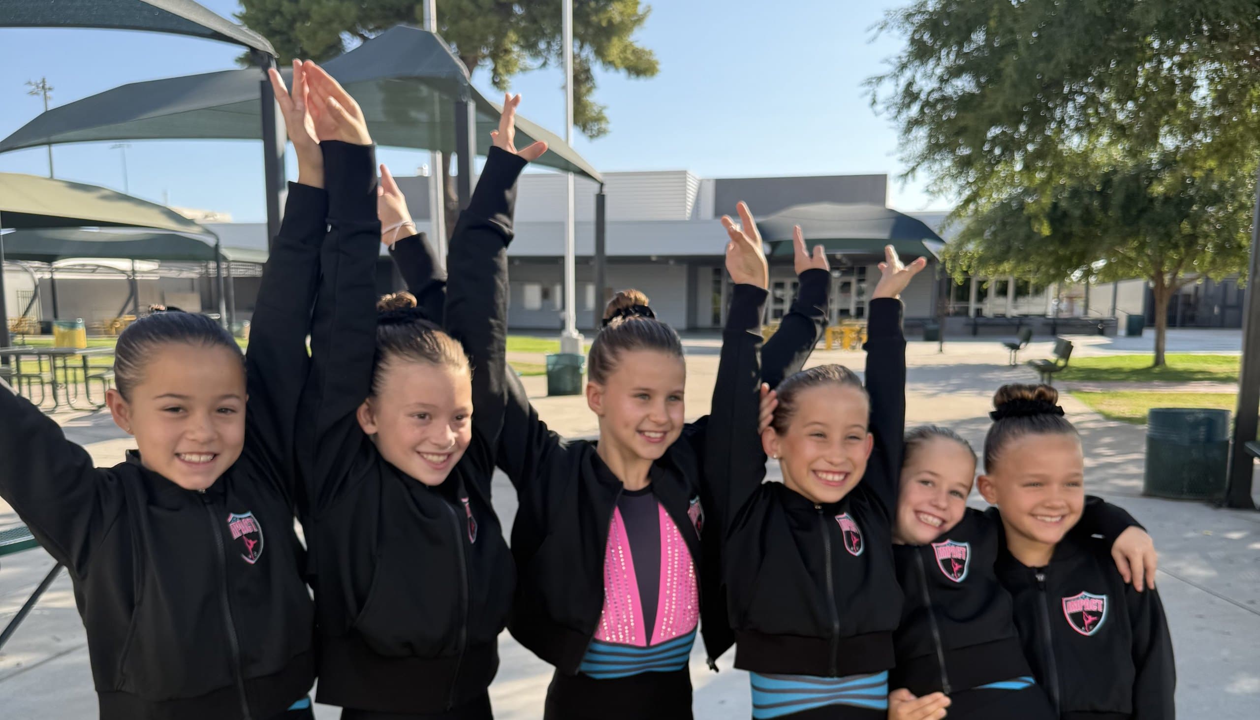 Impact Gymnastics team athletes in black team jackets with the Impact shield, arms raised and smiling